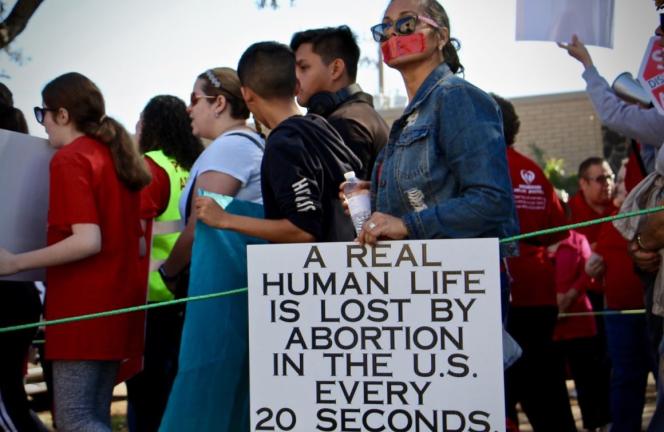 A person wearing a mask holding a sign that says "a human life is lost by abortion in the U.S every 20 seconds."