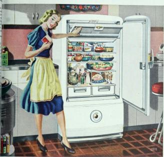 A white woman in a blue dress and a yellow apron, standing in front of an open fridge full of food.