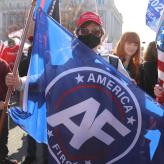 A man holding an "America First" flag, wearing a red MAGA hat, and a black face mask in a crowd of other right-wingers.