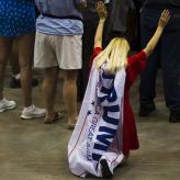 A white woman with blond hair, kneeling with her arms in the air, a Trump flag tied around her neck and hanging down her back
