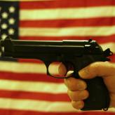 A hand gripping a handgun in front of an American flag backdrop