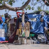 Boxes, prams, tarps, and other disparate items piled up under a blue sky on the sidewalk