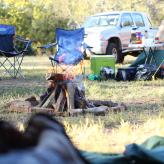 Stock photo of a camp with fire, camping chairs, the side of a tent, and a silver pickup truck in the background