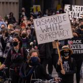 A person wearing a mask at a protest holding a sign that reads "Dismantle White Supremacy"