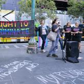 Protesters standing around a mic, two people holding a banner that reads "no right to discriminate"