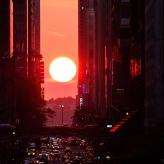 A sunset between buildings and a road full of vehicles.