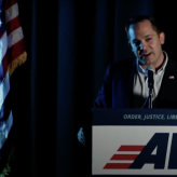 A man in blue suit standing at a podium with the letters AV on it speaking into a mic with two American flags behind him.