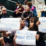 People sitting on stairs. Three people hold signs in Portugese, written with blue ink on white paper.