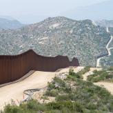 a brown fence following around a dirt road that fades into green mountains.