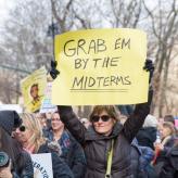A White woman in a black coat holding a banner saying "Grab em by the midterms"