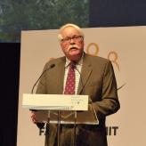 A man with white hair and a white mustache, speaking into a microphone.
