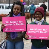 Three Black women wearing winter clothes holding signs that say "we march for Women's Health"