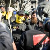 A Proud Boy with a neck warmer covering his mouth points mid-speech at the camera