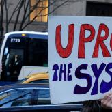 A person holds up a sign that reads "Uproot the system"
