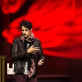 A man in a black jacket speaks in front of a red blurred background