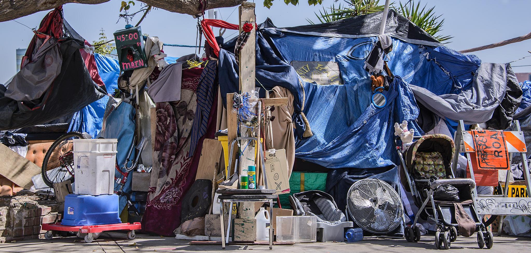 Boxes, prams, tarps, and other disparate items piled up under a blue sky on the sidewalk