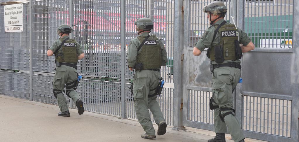 Three men in army training suits running along a large metal fence