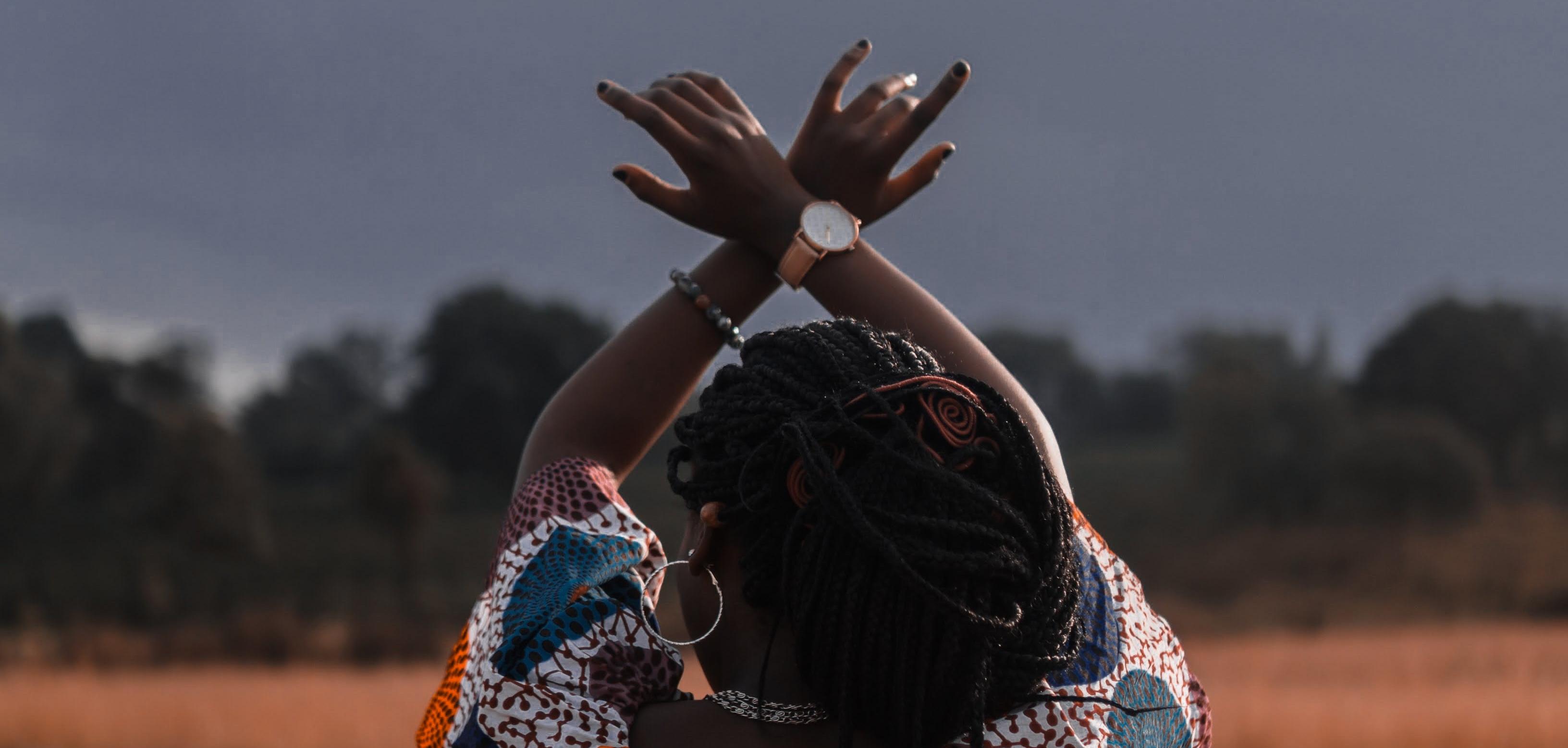 Black woman in field facing away from camera with arms in the air crossed at the wrist.