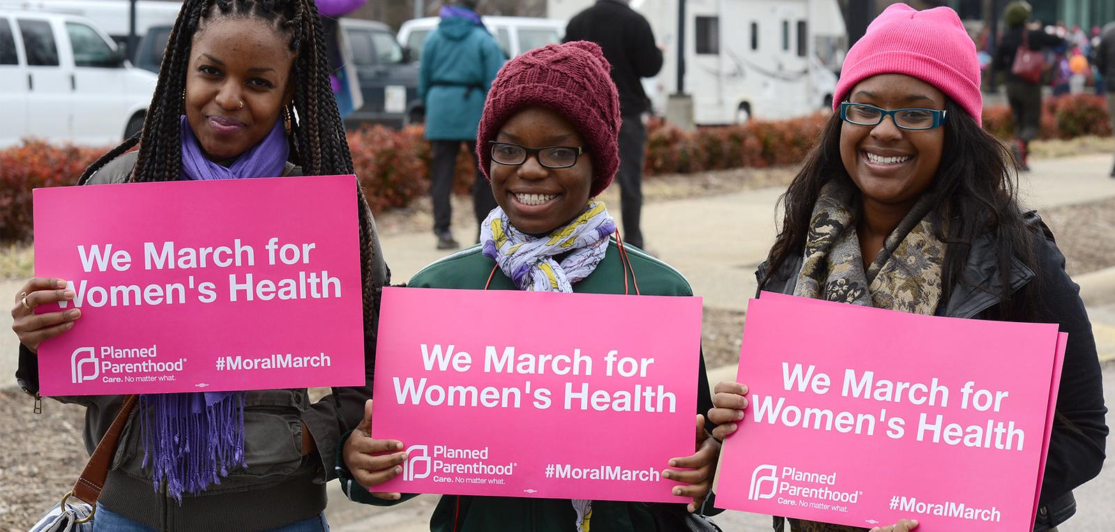 Three women holding signs that say "We march for Woomen's Health."