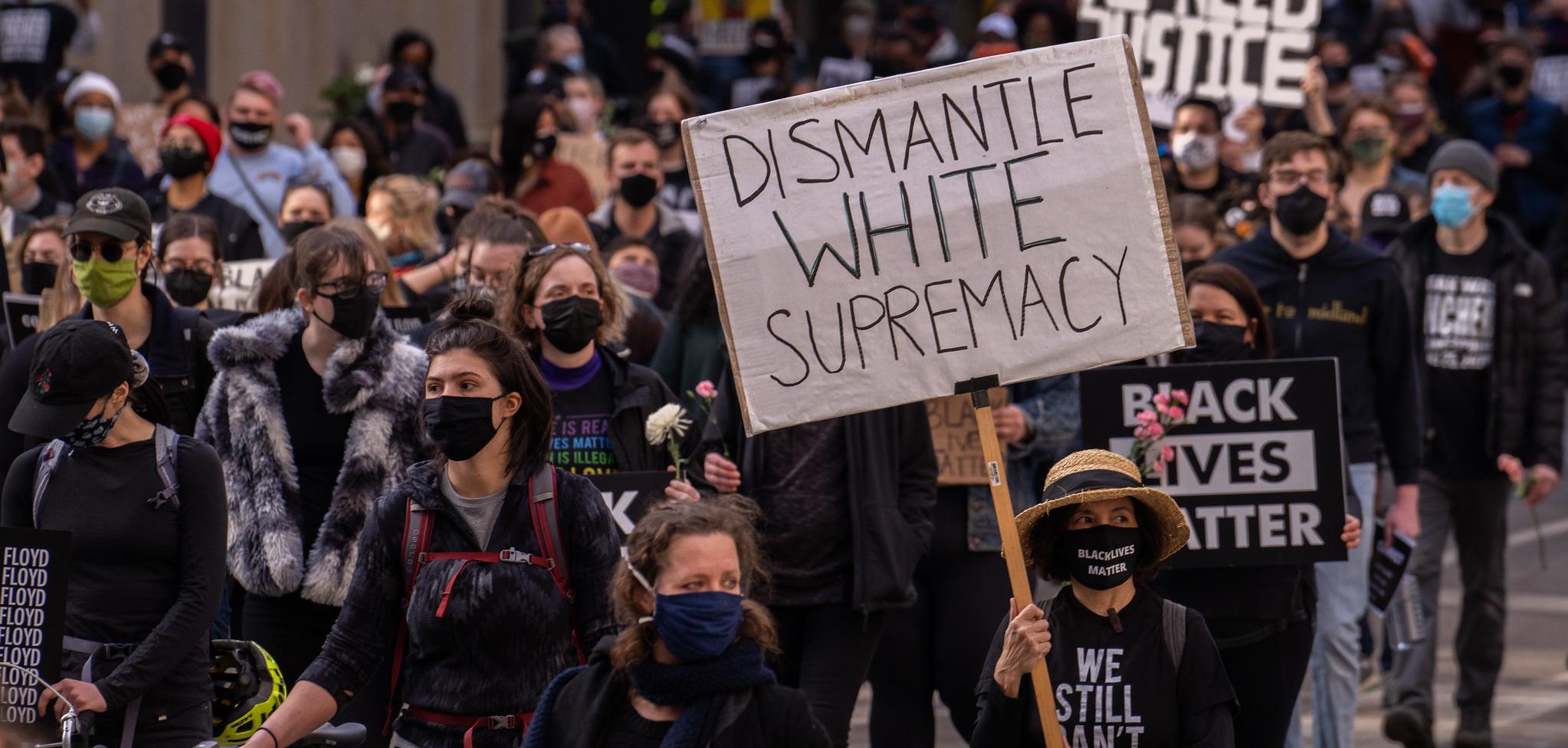 A person wearing a mask at a protest holding a sign that reads "Dismantle White Supremacy"