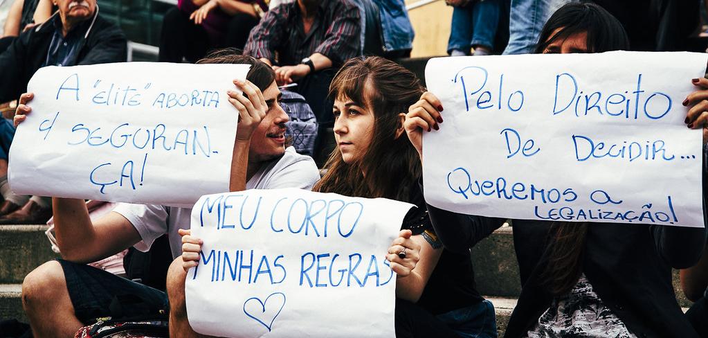 People sitting on stairs. Three people hold signs in Portugese, written with blue ink on white paper.