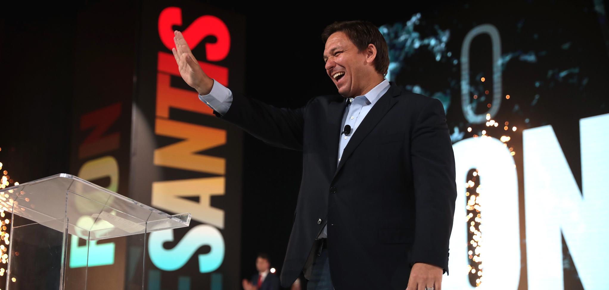 White man on stage with hand raised smiling, you can see the letters "ESANTIS" projected behind him.