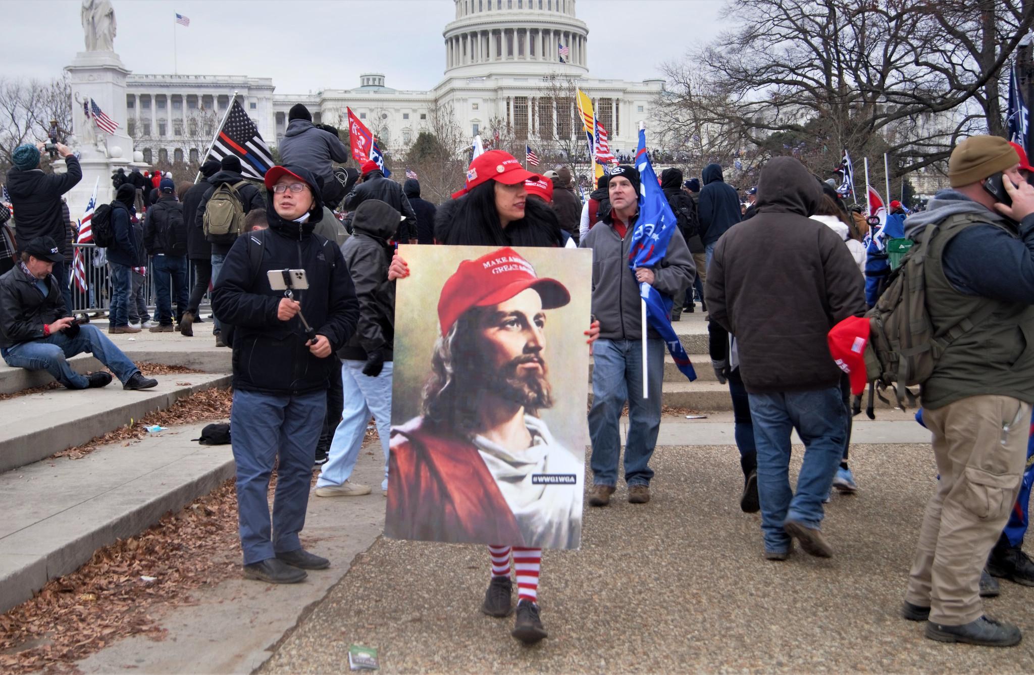 A woman wearing a Make America Great Again cap carrying an image of Jesus wearing a MAGA hat with "WWG1WGA" on his lapel