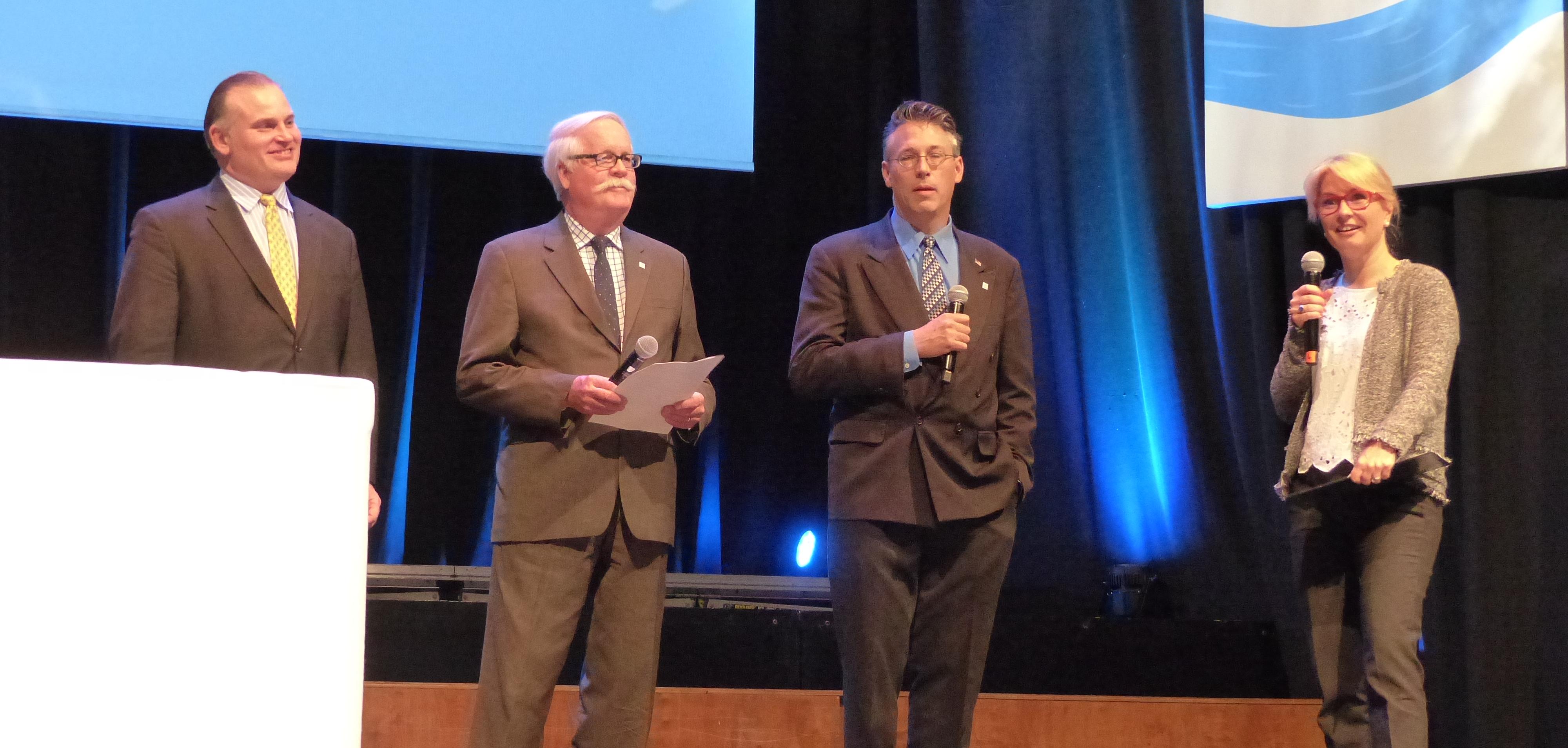 Three men in suits and a woman holding a mic.